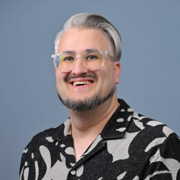 Portrait of Sean - a smiling young man with grey hair wearing glasses and a black and white geometric pattern shirt.