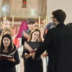 A close up shot of some members of the choir in rehearsal with the conductor with his back to the camera.