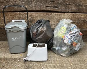 A toaster next to a grey food recycling caddy and a black sack of rubbish and a clear sack of recycling.