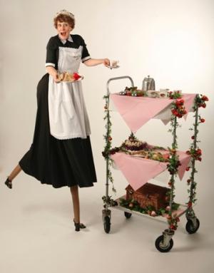 A woman on stilts dressed in a waitress outfit pushing a 3 tier trolly laden with cakes.