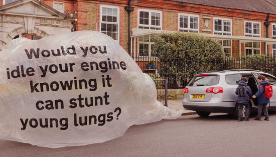Image of a car with 2 children outside a school. The car's exhaust pipe is inflating a huge bag with it's exhaust fumes. There is a message on the bag saying 'Would you idle your engine knowing it can stunt young lungs?'