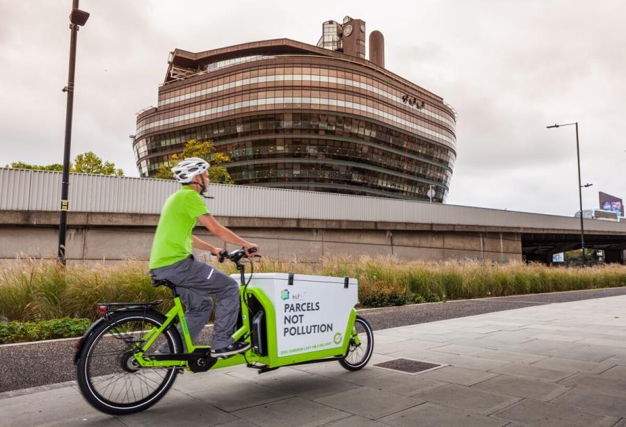 A man in a green t-shitt and cycle helmet riding a lime gree delivery bike with a large box at the front.