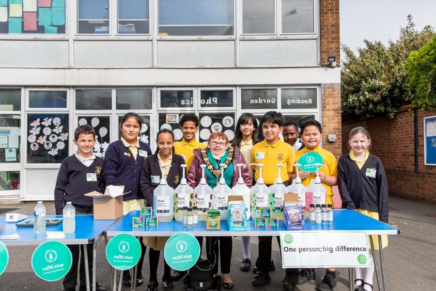 •	Refill Station - St John XXIII: A group of children and the Mayor of Hammersmith and Fulham Council stand in a playground in front of a table with refillable bottles and containers.