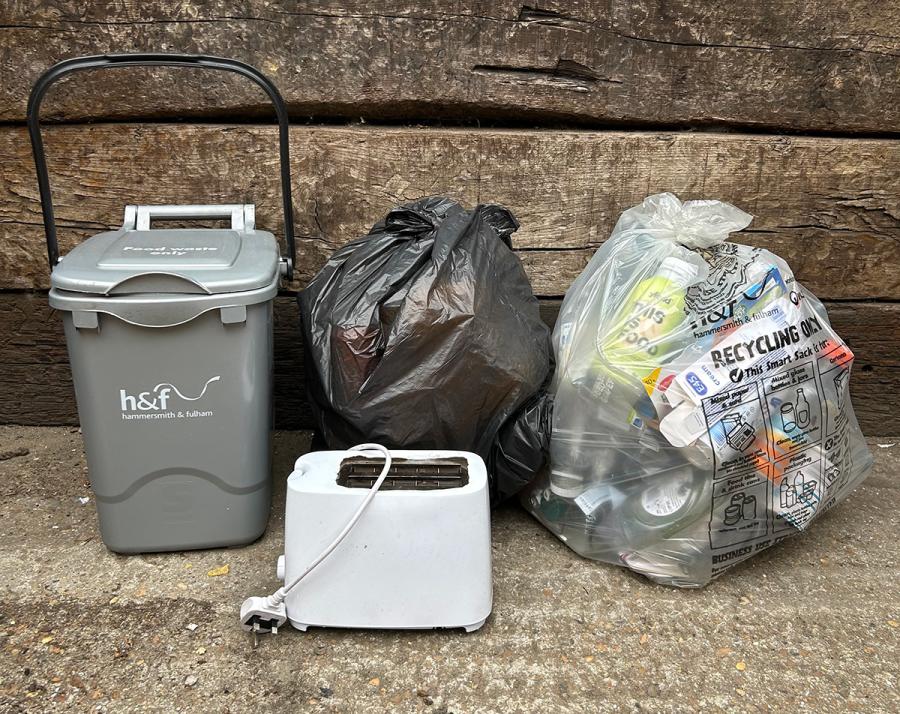 A toaster next to a grey food recycling caddy and a black sack of rubbish and a clear sack of recycling.