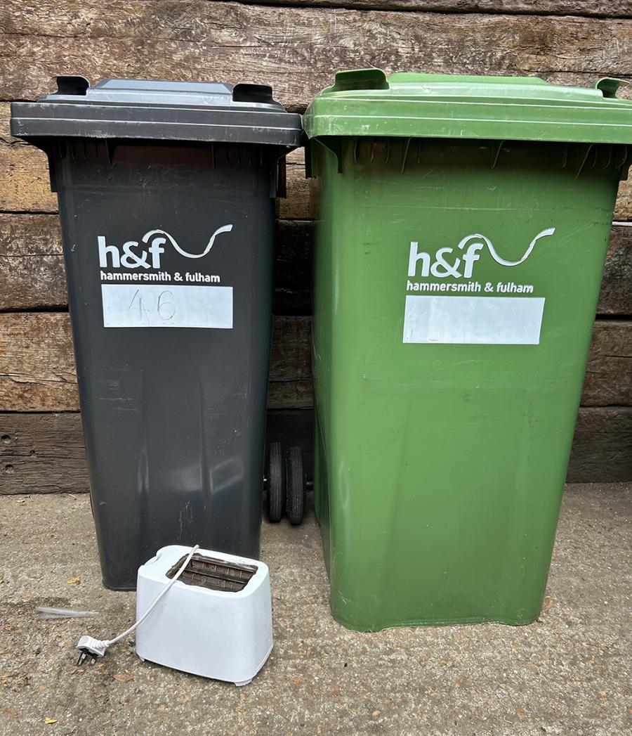 A toaster next to a black wheeled bin and a green wheeled bin.