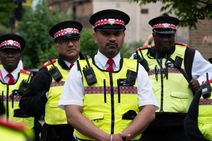A group of LET officers in their uniform which consists of black trousers, white shirts, red tie, high vis vest with reflective grey and red checked band and hat with red and white checked band.