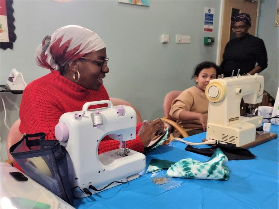 A woman and a girl operating sewing machines at a table while chatting to each other